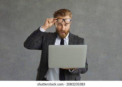 Oh my god, incredible. Excited surprised shocked man takes off glasses looking at laptop computer screen isolated on grey background. Lucky businessman makes deal or gets business email with good news