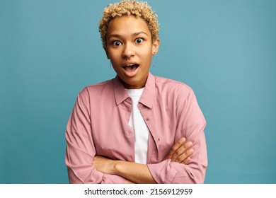 Oh my God! Shocked surprised and astonished female with blonde short afro hairstyle and dark skin standing against blue wall with opened mouth and big eyes after hearing unbelievable jaw dropping news