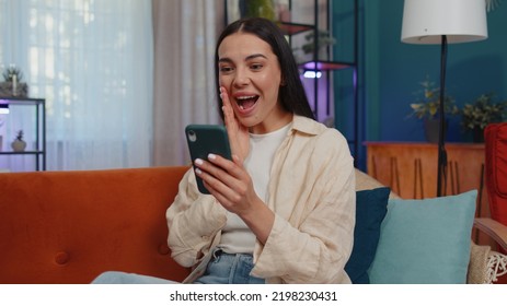 Oh My God Wow. Excited Happy Joyful Winner Girl Use Smartphone Typing Browsing Found Out Great Win Good News Celebrate Victory. Young Woman With Mobile Phone Lying At Home In Room On Orange Couch