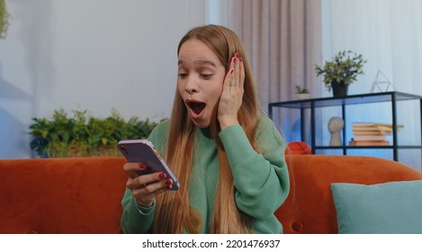 Oh My God Wow. Excited Happy Joyful Winner Girl Use Smartphone Typing Browsing Found Out Great Win Good News Celebrate Victory. Young Child Kid With Mobile Phone Sits At Home In Room On Orange Couch