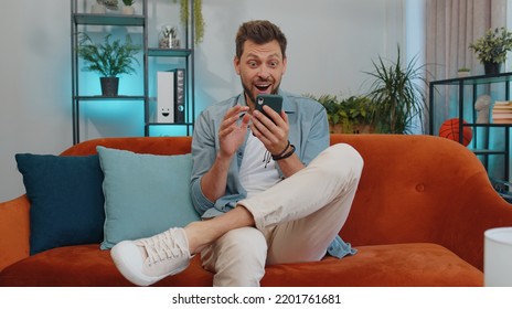 Oh My God Wow. Excited Happy Joyful Winner Man Use Smartphone Typing Browsing Found Out Great Win Good News Celebrate Victory. Young Guy With Mobile Phone Sitting At Home In Room On Orange Couch