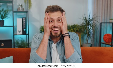 Oh My God, Wow. Handsome Adult Man Surprised Looking At Camera With Big Eyes, Shocked By Sudden Victory, Good Win News, Celebrating At Home. Excited Amazed Young Guy Indoors In Living Room On Couch