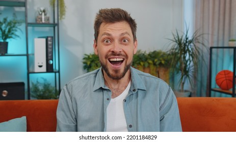 Oh My God, Wow. Handsome Adult Man Surprised Looking At Camera With Big Eyes, Shocked By Sudden Victory, Good Win News, Celebrating At Home. Excited Amazed Young Guy Indoors In Living Room On Couch