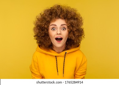 Oh my god, wow! Portrait of surprised curly-haired woman in urban style hoodie looking at camera with mouth open in amazement, expressing shock, astonishment. indoor studio shot, yellow background