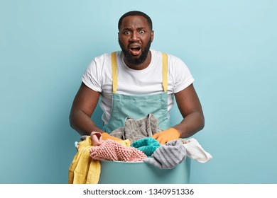 Oh No, Again Washing? Frustrated Black Laundry Hotel Worker Looks At Pile Of Clothes For Ironing, Opens Mouth Widely, Dressed In White T Shirt And Apron, Does Housework And Chores, Stands Indoor