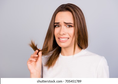 Oh No! Close Up Portrait Of Frustrated Young Brown Haired Woman Holding Her Hair Wih Separated Dry Ends, Standing On A Light Grey Background