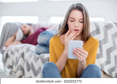 Oh No. Pretty Perplexed Long-haired Young Woman Feeling Surprised And Holding Her Mans Phone And He Lying On The Sofa