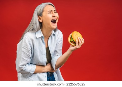 Oh, So Strong Pain In Stomach! Mature Senior Asian Woman Holding Burger In Hands. Stop Sign To Unhealthy Food, Isolated Over Red Background.