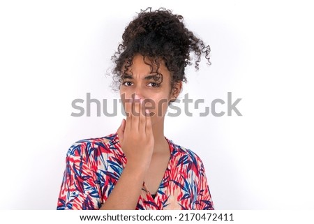 Oh! I think I said it! Close up portrait young beautiful brunette woman wearing colourful dress over white wall cover open mouth by hand palm, look at camera with big eyes.