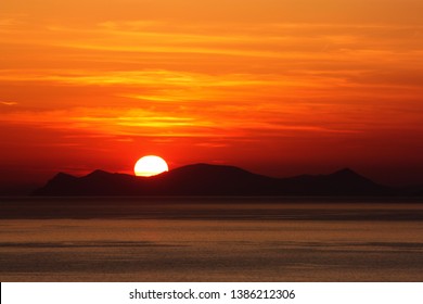 Oia santorini island greece. Natural coloured sunset. Dark red sky. Gold setting sun behind mountains. Calm sea reflecting sunlight. Beautiful colours. Thin clouds slightly visible. Mauntains - Powered by Shutterstock
