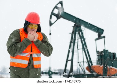 Oil Drill Field , Pump Jack In The Background .Oil And Gas Industry Worker In Safety Clothing Get It Frozen In Front , Blowing Hot Air In His Clasped Hands