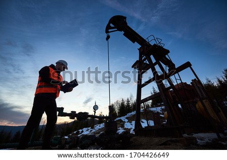 Oil engineer in work vest at oil pump rocker-machine, making notes while checking work of balanced beam petroleum pump jack under beautiful evening sky. Concept of oil extraction, petroleum industry.