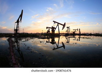 Ölfeld, am Abend, Ölpumpen laufen, die Ölpumpe und der schöne Sonnenuntergang reflektiert im Wasser, die Silhouette der Strahl Pump-Einheit am Abend.