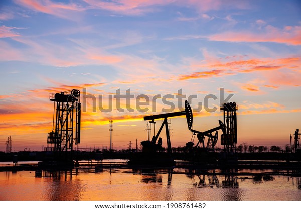 Oil field site, in the\
evening, oil pumps are running, The oil pump and the beautiful\
sunset reflected in the water, the silhouette of the beam pumping\
unit in the evening.