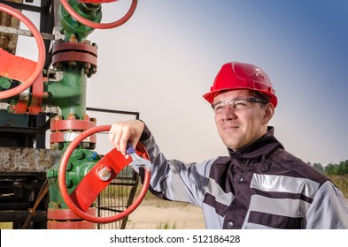 Oil Field Worker Near Wellhead Valve, Wearing Red Helmet And Work Clothes Holding Wrench In His Hands. Oil And Gas Concept.