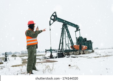 Oil And Gas Refinery Worker In Safety Clothing Pointing At Pump Jack In Oil Field And Talking On The Phone. Winter Season 