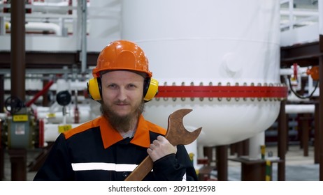 An Oil And Gas Worker Stands In A Hard Hat And Ear Protectors In A Gas Liquefaction Shop, Behind Him Is A Container Of Gas, And In His Hands Is A Large Wrench. Workplace Repairman.