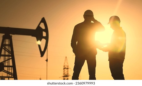 oil production. two silhouette workers work as a team next to an oil pump. business oil production production concept. two engineers of the oil and gas industry are sun discussing business plan - Powered by Shutterstock