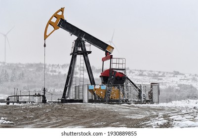 Oil Pump. Oil And Gas Industry Equipment. Oil Field Pump Jack And Oil Refinery In The Winter With Snow, Wind Turbine Mountains And Forest In Background