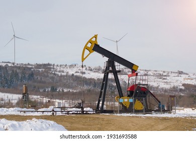 Oil Pump. Oil And Gas Industry Equipment. Oil Field Pump Jack And Oil Refinery In The Winter With Snow, Wind Turbine Mountains And Forest In Background