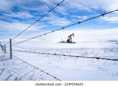 An Oil Pump Jack Working In A Snow Covered Agriculture Field Along A Barbed Wire Fence On The Canadian Prairies In Rocky View County Alberta Canada.