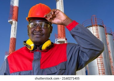 Oil Refinery Worker in Personal Protective Equipment Looking At Camera. Young African-American Oil Worker in Red Hardhat Standing In Front Of Oil Industry Storage Tanks.
