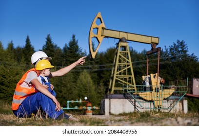 Oil Rig Operator Introducing His Son To His Profession. Oilman Squatting On Lawn With Male Child, Not Far From The Petroleum Well And Pointing Finger To Pump Jack.