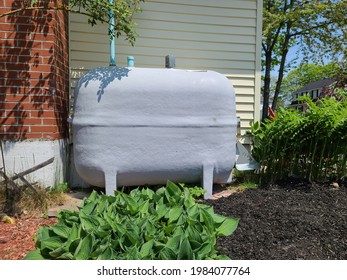 An Oil Tank Hidden On The Side Of A Suburban House In A Close Knit Community. The Tank Is Painted Gray And Nestled Next To A Brick Chimney And Flower Beds Filled With Hosta Plants.