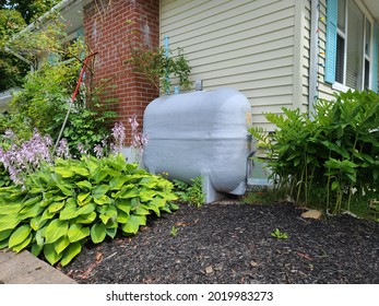 An Oil Tank Hidden On The Side Of A Suburban House In A Close Knit Community. The Tank Is Painted Gray And Nestled Next To A Brick Chimney And Flower Beds Filled With Hosta Plants.