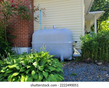 An Oil Tank Hidden On The Side Of A Suburban House In A Close Knit Community. The Tank Is Painted Gray And Nestled Next To A Brick Chimney And Flower Beds Filled With Hosta Plants.