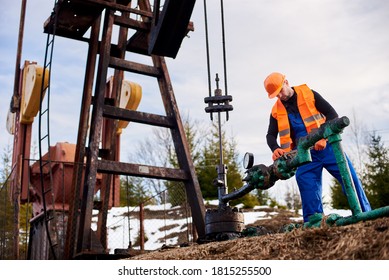 Oil Worker In Blue Overalls, Orange Vest And Helmet Using Industrial Wrench While Repairing Pipe Of Oil Well Pump Jack, Tightening Bolt On Pipe. Concept Of Petroleum Industry And Repair.