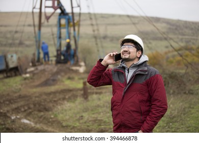 Oil Worker In Uniform And Helmet, With Mobile Phone On Of Background The Pump Jack And Sky. Toned. 