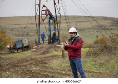 Oil Worker In Uniform And Helmet, With Mobile Phone On Of Background The Pump Jack And Sky. Toned. 