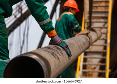 Oil Workers In Green And Orange Workwear And Work Gloves On Oil Deposit. With Rusted Drilling Pipe. Focus On Foreground On First Worker's Hand In Glove. Kazakhstan