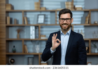 OK sign, businessman shows approval sign to camera, smiling man in business suit inside office at workplace, experienced financier investor in office.