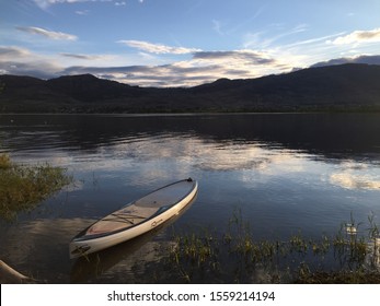 Okanagan Lake, Canada; June 14, 2017- Empty Paddle Board Floating On The Lake With Mountains And Sunset In The Background And Long Grass In The Foreground. Concept Image For Serenity And Relaxation