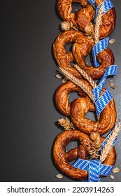 Oktoberfest concept - pretzels with traditional pattern ribbon and ears of wheat. A trendy hard light, dark shadow, black stone concrete background, flat lay, top view