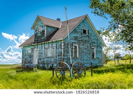 Old, abandoned blue prairie farmhouse with trees, grass, blue sky and a wagon wheel in Kayville, Saskatchewan, Canada