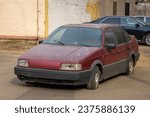 An old abandoned broken car with flat tires on the city parking. The scrap and recycling vehicle. The dusty and rusty machine body and the broken headlight