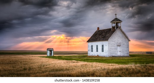 Old abandoned church in the countryside at sunset. Sun rays are beaming down on the church from the clouds above. There is an old outhouse visible off to the side of the church. 