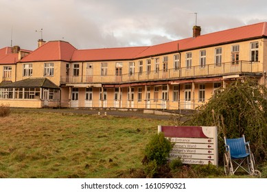  Old Abandoned Nursing Home, Hospital Building Exterior, Delapelated With Old Wheelchair Abandoned In Foreground Beside Bush, With Broken Windows And Doors Boarded Up.