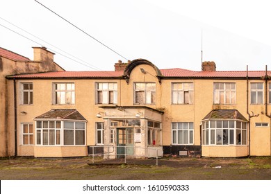 Old Abandoned Nursing Home, Hospital Building Exterior Front, Delapelated Front Entrance Porch In The Foreground, And Broken Windows Boarded Up And Doors Open.