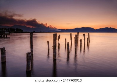 Old abandoned pier in shallow coast water in the orange morning light with a smoking forest fire in the hills in the background - Powered by Shutterstock