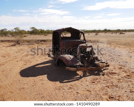 An old abandoned rusted out shell of an old ute truck slowly rotting and rusting on the red dirt of the desert with a line of green trees along a dried up river bed and blue sky on Central Australia