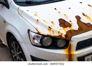 An old abandoned rusty white car with damaged paintwork on the bumper and hood. 