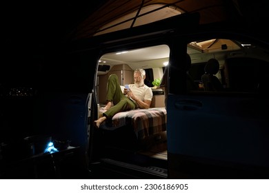 Old active man sitting inside rv camper van with tent on it on vacation using mobile phone. Senior mature traveler holding smartphone enjoying free internet or checking rentals in camping at night. - Powered by Shutterstock