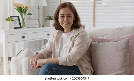 Old Adult Asia Curly Hair Female Sitting Easy Relax On Sofa Couch Toothy Smile Looking At Camera At Cozy Home For Happy Early Retired In Older People, Elderly Mental Health Care, Aging Skin Lady Face.