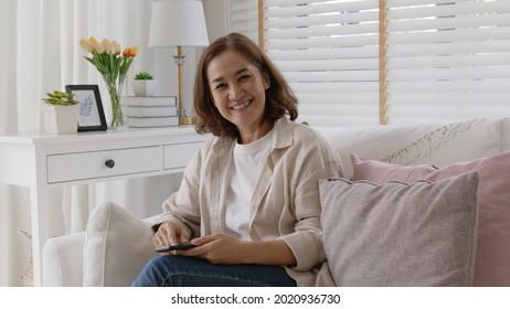 Old adult asia curly hair female sitting easy relax on sofa couch toothy smile looking at camera at cozy home for happy early retired in older people, elderly mental health care, aging skin lady face.