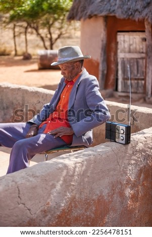 old african village man wearing a hat listening to radio receiver in his yard