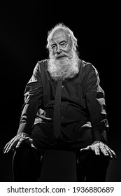 Old age concept. Black-and-white portrait of an old man with white beard sits and smiles at camera. Black background. 